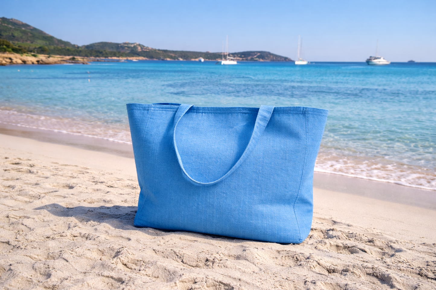 Blue tote bag on a sandy beach with clear blue water and mountains in the background