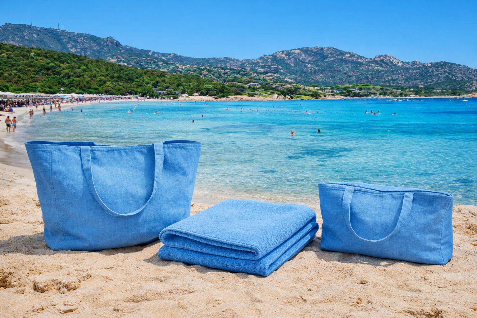 Two blue beach bags and a blue towel on a sandy beach with mountains and clear blue water in the background.