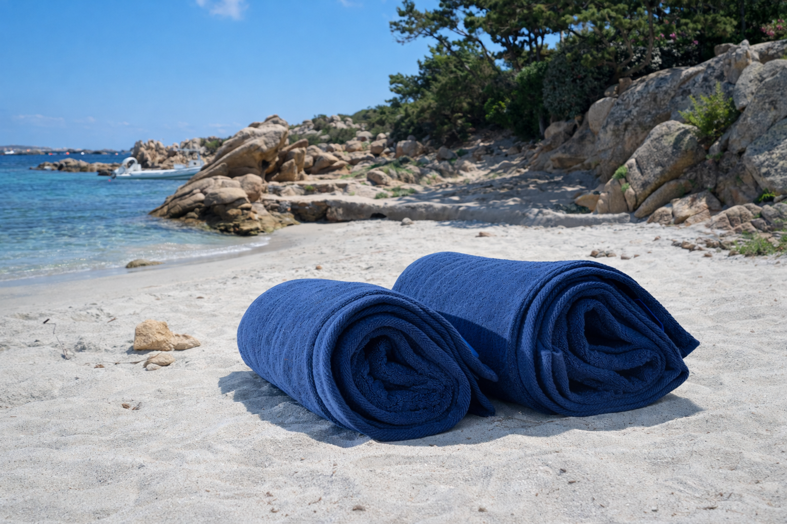 Two rolled-up blue towels on a sandy beach with rocky cliffs and water in the background.