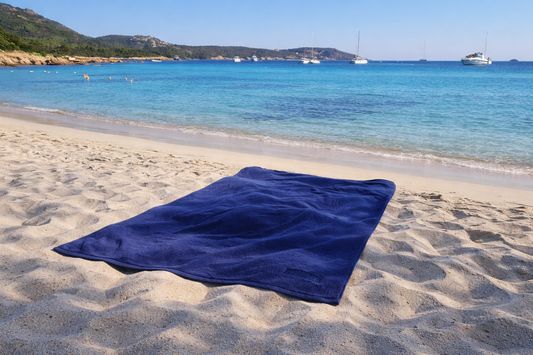 Blue towel on a sandy beach with clear blue water and boats in the background