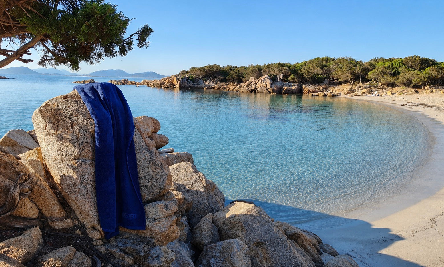 A vibrant royal blue towel draped over a large rock on a sunny beach, overlooking clear turquoise water and a sandy cove with rocky shores and green trees.