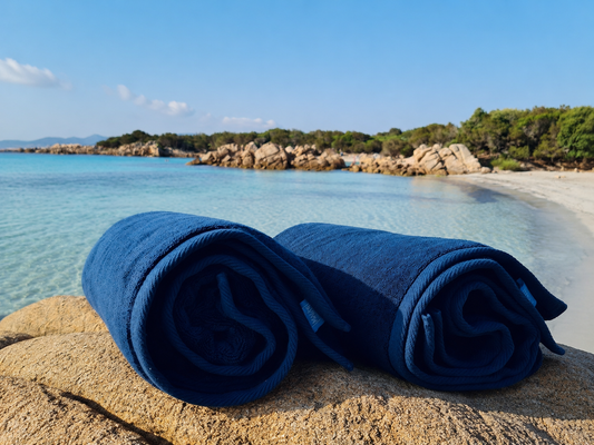 Two rolled dark blue towels resting on a sandy beach with turquoise water and rocky shoreline in the background.