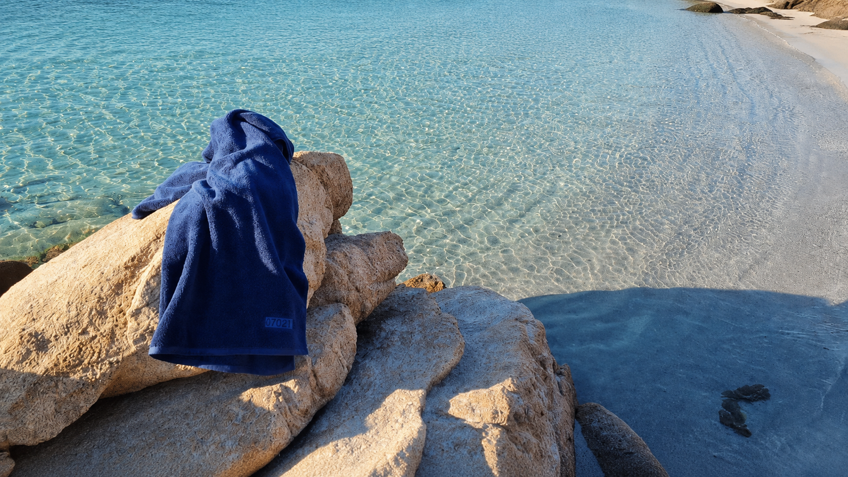 A dark blue hooded towel or beach wrap draped over a large tan rock, overlooking a sunny beach with clear turquoise water and pale sand.